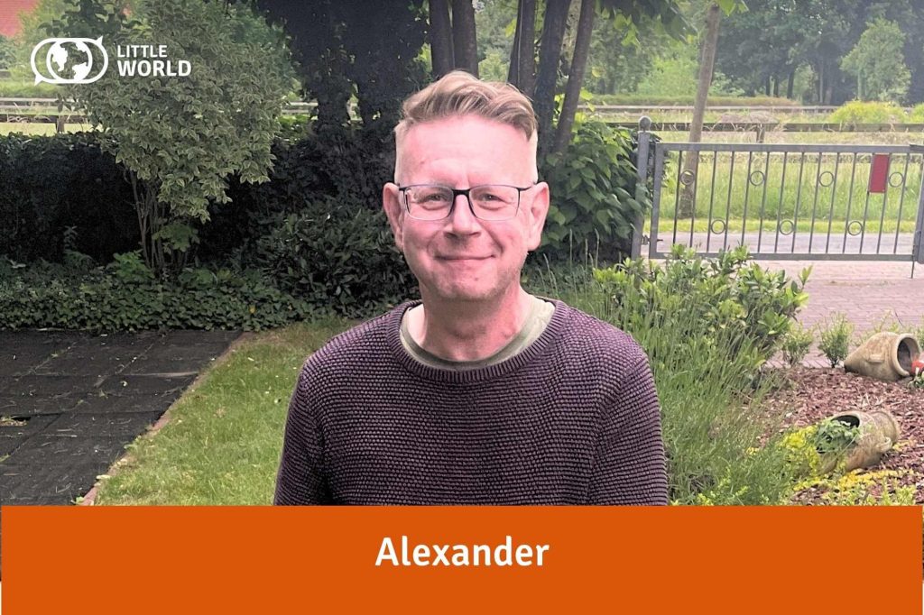 Alexander, ein Mann mit kurzen blonden Haaren und Brille steht lächelnd in einem Garten