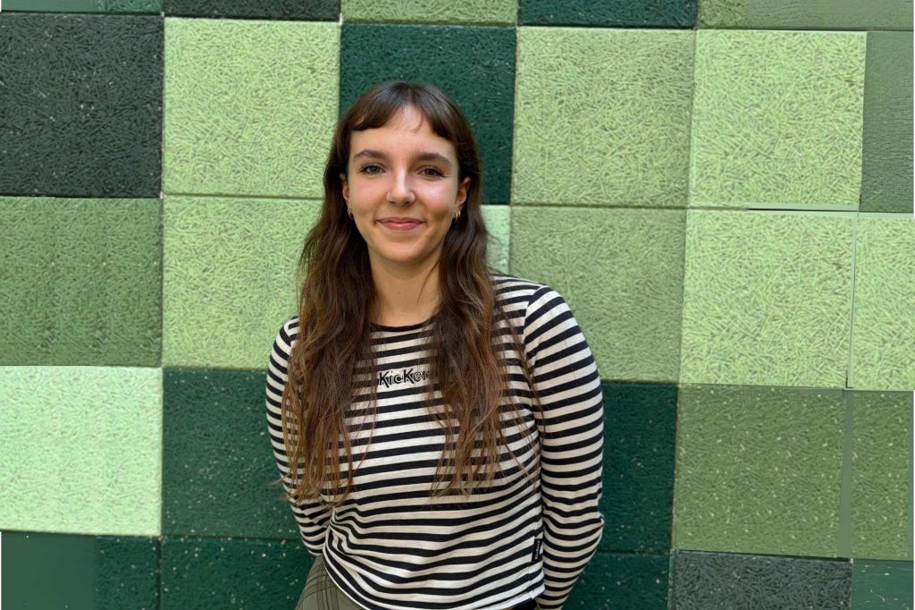 Alice, eine junge Frau mit langen brauen Haaren und Ringelshirt steht vor einer grün gekachelten Wand