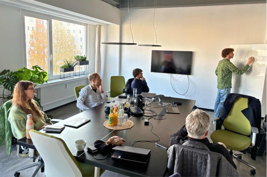 Workshopteilnehmer hören Tim zu, der etwas sm Whiteboard erklärt