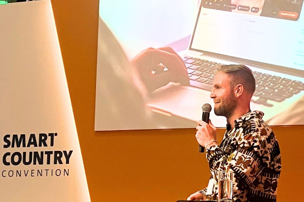 Oliver auf der Bühne der Smart Country Convention 2025. Großaufnahme von Oliver bei seinem Vortrag, hinter ihm sieht man eine Aufnahme des Little World Videochats auf der Leinwand