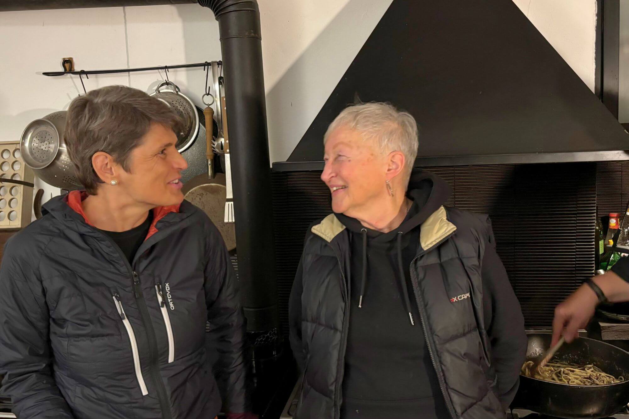 Susanne uns Sibylle in einer urigen Küche, im Hintergrund rührt jemand in einer Pfanne Pasta