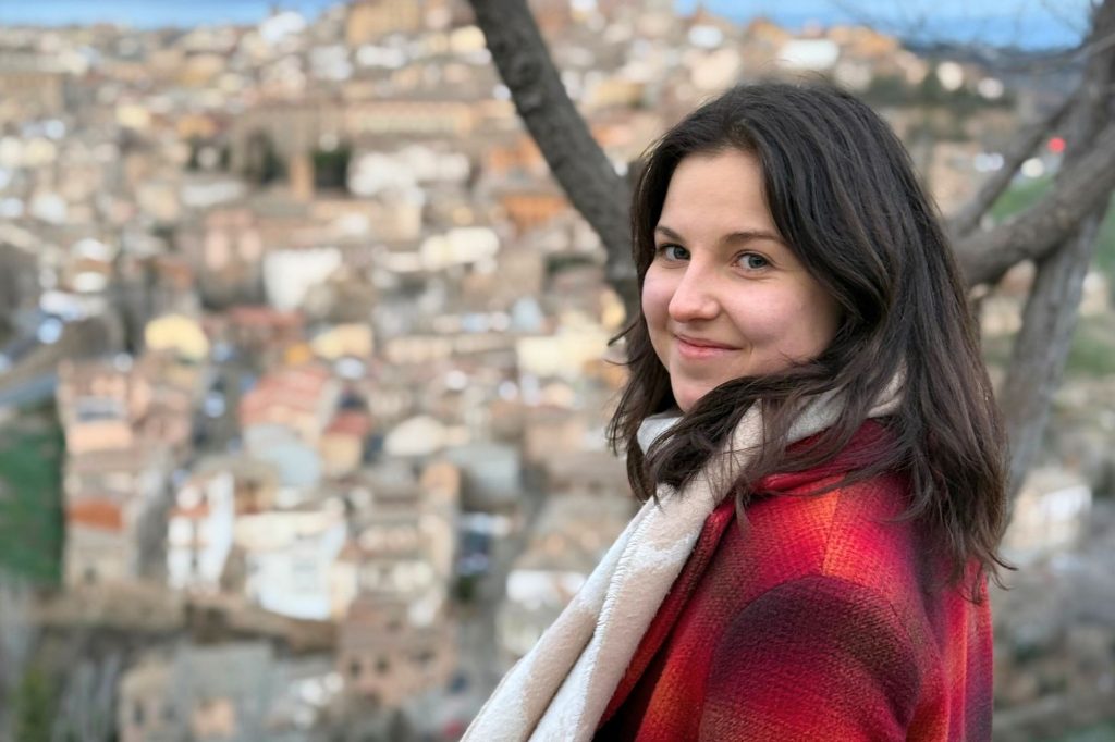 Junge Frau mit langen dunklen Haaren und rotem Mantel steht vor einem Baum auf einem Aussichtspunkt über einer Stadt