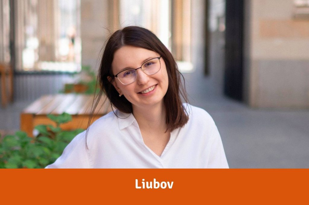 Junge Frau mit langen braunen Haaren, Brille und weißer Bluse sitzt in einem Hinterhof