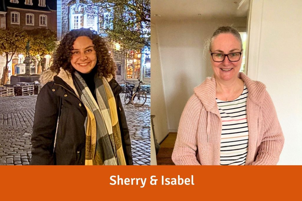 Sherry, eine junge Frau mit braunen Locken, und Isabel eine Frau mittleren Alters mit Brille und zurückgebundenen blonden Haaren, lächeln in die Kamera
