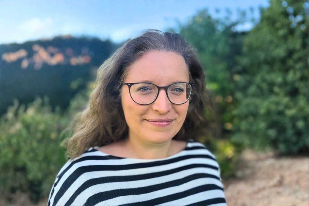 Sara, eine Frau mit blonden langen Haaren und Brille im gestreiften Shirt steht draußen vor einem Baum