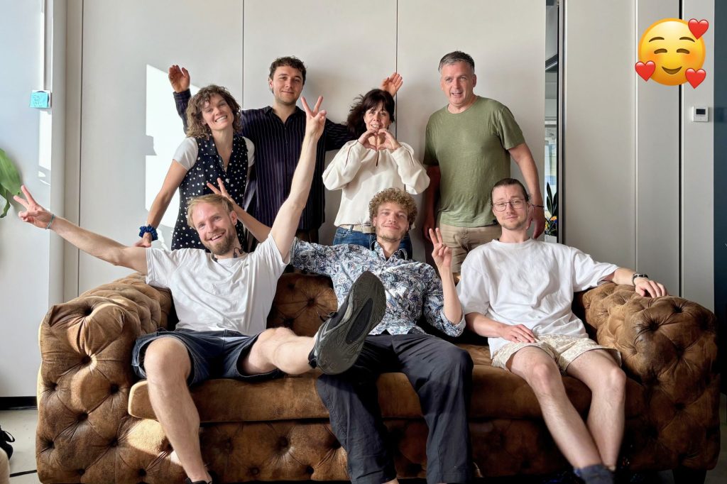 Team von Little World posiert fröhlich auf einem braunen Ledersofa mit Gesten wie Herzformen und Peace-Zeichen.